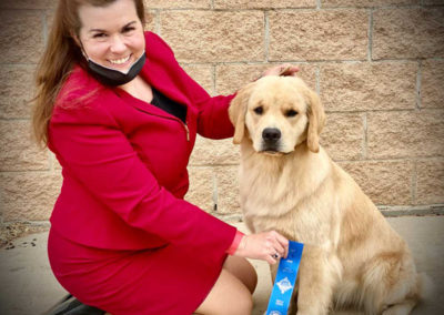 Woman in pink suit holding blue ribbon in front of a golden retriever puppy