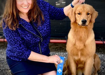 Young golden retriever sitting next to smiling woman holding a blue ribbon