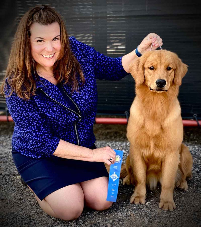 Young golden retriever sitting next to smiling woman holding a blue ribbon