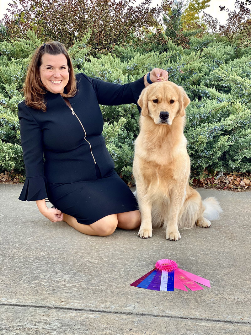 Woman in black suit with a beautiful golden retriever sitting behind multi-colored ribbons