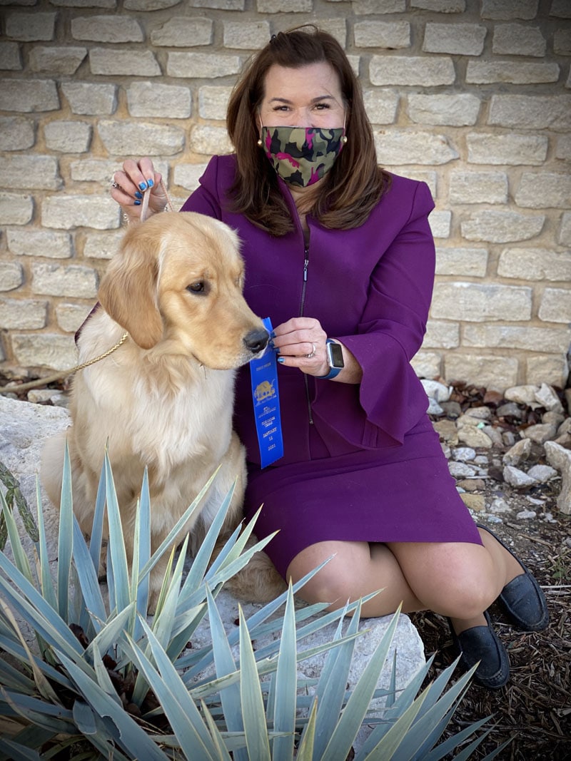 Woman in purple suit holding blue ribbon next to a golden retriever